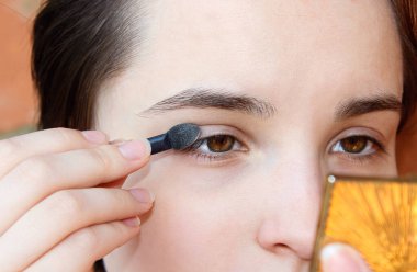 Genç bir kız aynanın önüne fırçayla göz kapaklarına gölgeler koyar. Kapat..