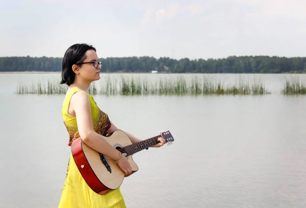 A girl in a dress plays the guitar standing barefoot in the water of a lake in nature. The vertical frame.