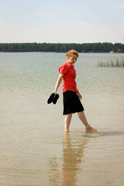 A young businesswoman in office clothes walks barefoot on the water on a lake in the heat and holds high-heeled shoes in her hands. Freelancing, the concept of leisureRear view.