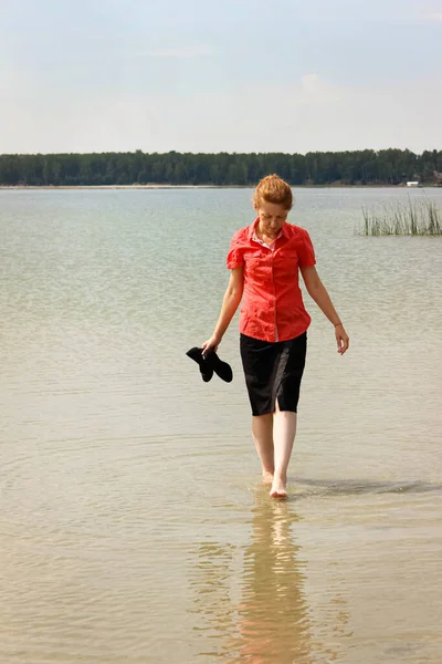 A young businesswoman in office clothes walks barefoot on the water on a lake in the heat and holds high-heeled shoes in her hands. Freelancing, the concept of leisureRear view.