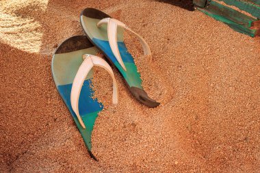 Altın sarısı bir kumsalda, ahşap mobilyaların olduğu bir barda renkli parmak arası terlikler.