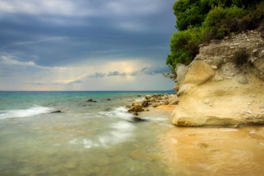 Manzaralı Kalogria plajı güneş ışınları bulutlardan ve ön plandaki büyük kayalardan geçiyor.