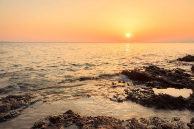 Yansıtıcı deniz suyunda aşınmış kaya tabakaları ve ufukta batan güneş.