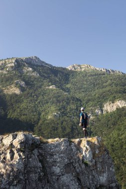 Sırt çantası, şapkası ve akıllı saati olan dağ yürüyüşçüsü kayalık bir uçurumun kenarında duruyor ve etkileyici dağ zirvelerine bakıyor.