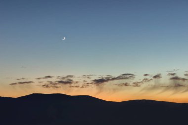Genç ay, dağ siluetleri ve güzel gün batımı gökyüzü alçak kabarık bulutlarla