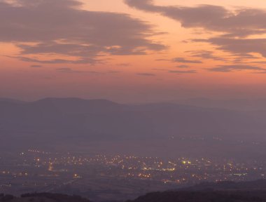 Uzak şehrin üzerindeki dramatik, renkli gün batımı. Sis ve siluet ufuk dağlarıyla kaplı.