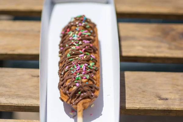 A waffle on a stick, covered with chocolate and colorful candies, served in a paper box