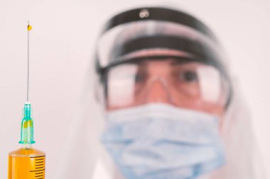 Maskeli ve koruyucu gözlüklü bir laboratuar bilimcisi elinde sarı bir sıvıyla bir şırınga tutuyor. Sığ derinlik alanı ile iğne tepesinde düşüşe odaklan
