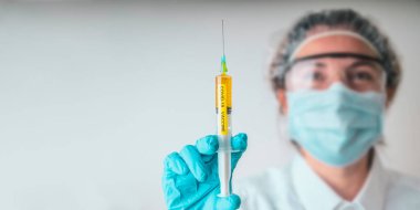 Maskeli ve koruyucu gözlüklü kadın bir laboratuvar bilimcisi COVID-19 aşısı olan bir şırınga tutuyor.