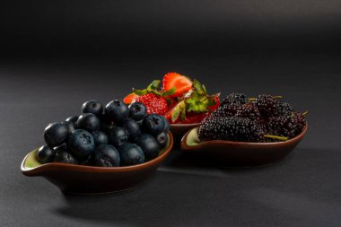 Önündeki doğal ve organik böğürtlenleri yakından çek. Ortada siyah dutlar ve bulanık kırmızı çilekler. Koyu arkaplanda taze meyve ve sığ alan derinliği