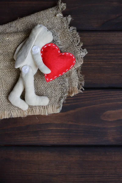Red heart in the hands of a cute hare. Soft toy rabbit holds a felt symbol of love with decorative white seams in its paws. Greeting card for Valentine\'s Day holiday with brown wooden texture