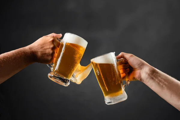 two hands with beer mugs, toasting in celebration, with overflowing foam, dark background and space for writing