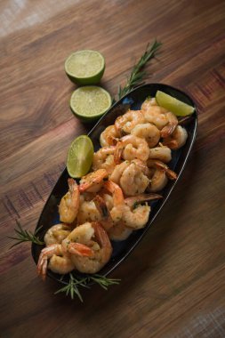 Sarımsak ve yağlı karides, ahşap bir masada koyu bir tabakta servis edilir, biberiye ve limonla süslenir.