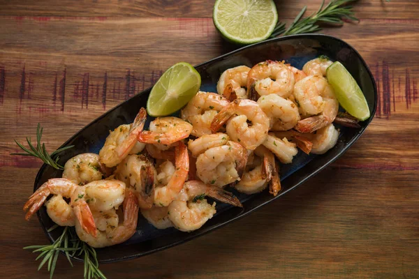 top view shrimp with garlic and oil, served on a dark plate on a rustic wooden table, decorated with rosemary and lemon