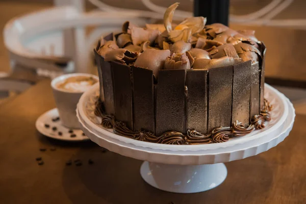 チョコレートケーキの上にダークチョコレートのシェービング ホワイトプレート 背景にコーヒーとカップ 木製のテーブルの選択的フォーカス — ストック写真