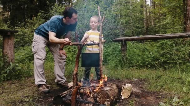 O pai ensina o filho a cozinhar na fogueira. Criança está cozinhando mingau de cereal em panela pendurada sobre o fogo na floresta. Acampamento turístico — Vídeo de Stock