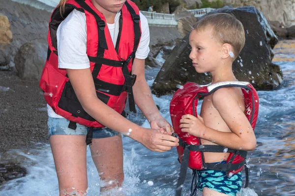 Girl lifeguard helps put on life vest on little boy. Mother dresses son in red jacket in waves. Child safety in open water