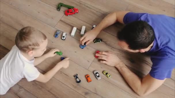 Padre joven e hijo pequeño se encuentran en el suelo de madera caliente de la casa y jugar en coches de juguete de colores. Infancia divertida, familia feliz. El niño conduce muchos coches. Mini transporte. Vista superior — Vídeos de Stock