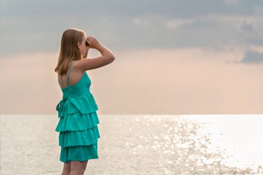 Denizde dürbünle bakan güzel bir kız. Uzun saçlı sarışın kadın ayakta duruyor ve gemilere bakıyor.