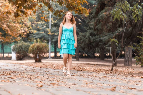 Girl in turquoise dress walks in autumn park with fallen yellow leaves. Autumn. Beautiful happy female with hair walking in city
