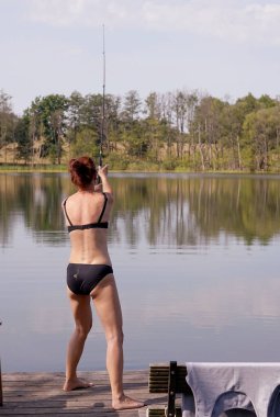 Bikinili bir kadın göldeki iskeleden olta atıyor.