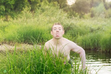 Soyunmuş genç bir adam çim çalılarından dışarı baktı ve dilini gösterdi..