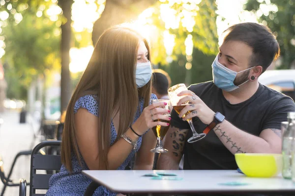 Spain Madrid. Caucasian couple toasting with beer wearing sanitary mask. Reopen pubs and club after quarentine coronavirus Covid-19. Man and woman drinking beer. Cheers. People smiling wearing mask