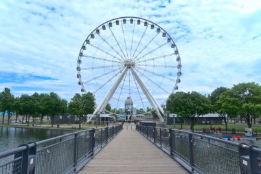 Montreal Grand Ferris Wheel, Old Port 'ta bir adada. Arka plan mavi bulutlu bir gökyüzü. Ön plan bir köprüdür.