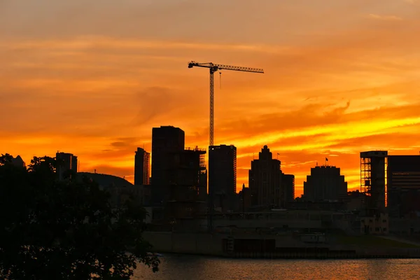 A skyline view of a city with a lifting crane in a construction site in the middle and a foreground a river, Montreal Downtown and Saint Laurent River