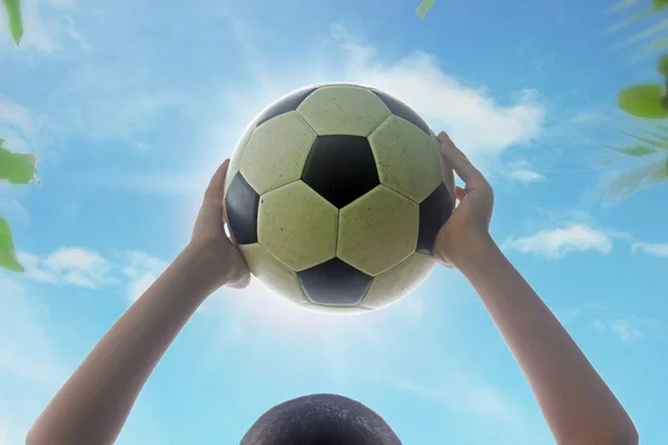 Çocuk futbol topunu başının üzerinde tutuyor. Gökyüzünün parlak ve parlak zemininde.