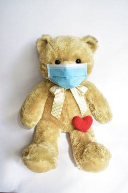 Beyaz arka planda mavi tıbbi maskeli sevimli kahverengi ayı, kırmızı kalp. Yüz maskesi takan oyuncak ayı Covid-19 Corona virüsü yayılmasını önlüyor.