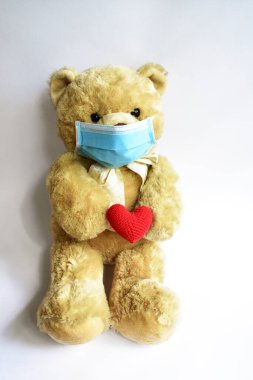 Beyaz arka planda mavi tıbbi maskeli sevimli kahverengi ayı, kırmızı kalp. Yüz maskesi takan oyuncak ayı virüs bulaşmasını önlüyor. CoV-2 Corona virüsü hastalığı karantina altında.
