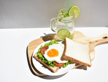 Sandviçli sağlıklı kahvaltı yemeği, kızarmış yumurta, domates dilimi, salata, nane yaprağı, mason kavanozu bardak limonata, ahşap kesim tahtası, beyaz arka plan, güneşli, abur cubur, kilo kaybı, organik, basitlik, vegan, burger, düşük yağ oranı, spor yemeği.