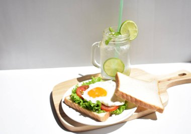 Sandviçli sağlıklı kahvaltı yemeği, kızarmış yumurta, domates dilimi, salata, nane yaprağı, mason kavanozu bardak limonata, ahşap kesim tahtası, beyaz arka plan, güneşli, abur cubur, kilo kaybı, organik, basitlik, vegan, burger, düşük yağ oranı, spor yemeği.