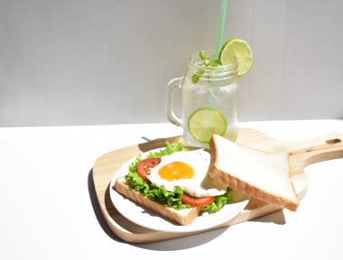 Sandviçli sağlıklı kahvaltı yemeği, kızarmış yumurta, domates dilimi, salata, nane yaprağı, mason kavanozu bardak limonata, ahşap kesim tahtası, beyaz arka plan, güneşli, abur cubur, kilo kaybı, organik, basitlik, vegan, burger, düşük yağ oranı, spor yemeği.