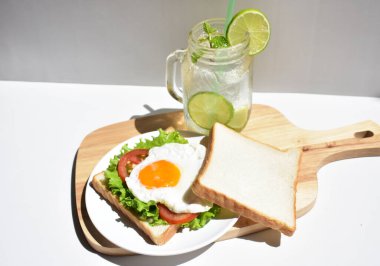 Sandviçli sağlıklı kahvaltı yemeği, kızarmış yumurta, domates dilimi, salata, nane yaprağı, mason kavanozu bardak limonata, ahşap kesim tahtası, beyaz arka plan, güneşli, abur cubur, kilo kaybı, organik, basitlik, vegan, burger, düşük yağ oranı, spor yemeği.
