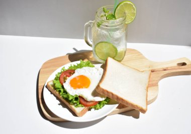 Sandviçli sağlıklı kahvaltı yemeği, kızarmış yumurta, domates dilimi, salata, nane yaprağı, mason kavanozu bardak limonata, ahşap kesim tahtası, beyaz arka plan, güneşli, abur cubur, kilo kaybı, organik, basitlik, vegan, burger, düşük yağ oranı, spor yemeği.