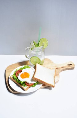 Sandviçli sağlıklı kahvaltı yemeği, kızarmış yumurta, domates dilimi, salata, nane yaprağı, mason kavanozu bardak limonata, ahşap kesim tahtası, beyaz arka plan, güneşli, abur cubur, kilo kaybı, organik, basitlik, vegan, burger, düşük yağ oranı, spor yemeği.