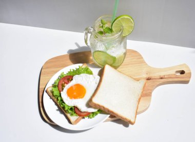 Sandviçli sağlıklı kahvaltı yemeği, kızarmış yumurta, domates dilimi, salata, nane yaprağı, mason kavanozu bardak limonata, ahşap kesim tahtası, beyaz arka plan, güneşli, abur cubur, kilo kaybı, organik, basitlik, vegan, burger, düşük yağ oranı, spor yemeği.