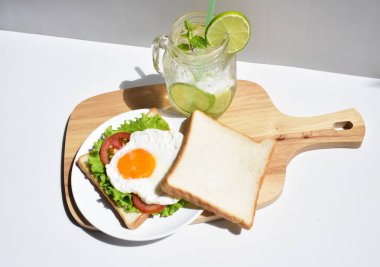 Sandviçli sağlıklı kahvaltı yemeği, kızarmış yumurta, domates dilimi, salata, nane yaprağı, mason kavanozu bardak limonata, ahşap kesim tahtası, beyaz arka plan, güneşli, abur cubur, kilo kaybı, organik, basitlik, vegan, burger, düşük yağ oranı, spor yemeği.