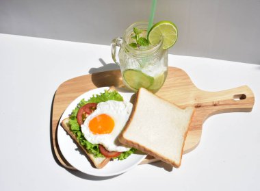 Sandviçli sağlıklı kahvaltı yemeği, kızarmış yumurta, domates dilimi, salata, nane yaprağı, mason kavanozu bardak limonata, ahşap kesim tahtası, beyaz arka plan, güneşli, abur cubur, kilo kaybı, organik, basitlik, vegan, burger, düşük yağ oranı, spor yemeği.