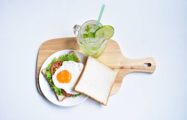 Sandviçli sağlıklı kahvaltı yemeği, kızarmış yumurta, domates dilimi, salata, nane yaprağı, mason kavanozu bardak limonata, ahşap kesim tahtası, beyaz arka plan, güneşli, abur cubur, kilo kaybı, organik, basitlik, vegan, burger, düşük yağ oranı, spor yemeği.