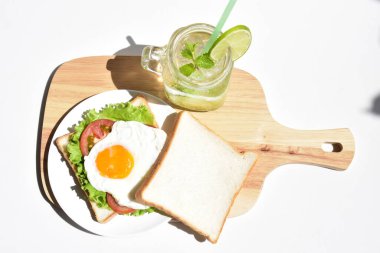 Sandviçli sağlıklı kahvaltı yemeği, kızarmış yumurta, domates dilimi, salata, nane yaprağı, mason kavanozu bardak limonata, ahşap kesim tahtası, beyaz arka plan, güneşli, abur cubur, kilo kaybı, organik, basitlik, vegan, burger, düşük yağ oranı, spor yemeği.
