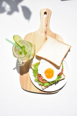 Sandviçli sağlıklı kahvaltı yemeği, kızarmış yumurta, domates dilimi, salata, nane yaprağı, mason kavanozu bardak limonata, ahşap kesim tahtası, beyaz arka plan, güneşli, abur cubur, kilo kaybı, organik, basitlik, vegan, burger, düşük yağ oranı, spor yemeği.