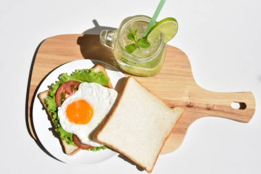 Sandviçli sağlıklı kahvaltı yemeği, kızarmış yumurta, domates dilimi, salata, nane yaprağı, mason kavanozu bardak limonata, ahşap kesim tahtası, beyaz arka plan, güneşli, abur cubur, kilo kaybı, organik, basitlik, vegan, burger, düşük yağ oranı, spor yemeği.