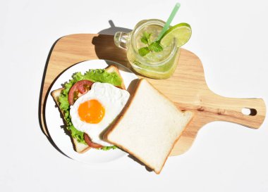 Sandviçli sağlıklı kahvaltı yemeği, kızarmış yumurta, domates dilimi, salata, nane yaprağı, mason kavanozu bardak limonata, ahşap kesim tahtası, beyaz arka plan, güneşli, abur cubur, kilo kaybı, organik, basitlik, vegan, burger, düşük yağ oranı, spor yemeği.