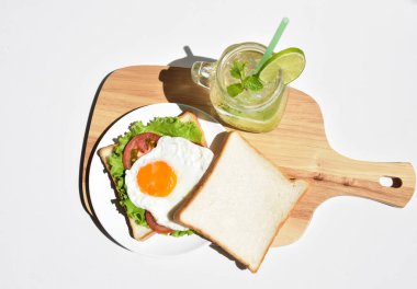 Sandviçli sağlıklı kahvaltı yemeği, kızarmış yumurta, domates dilimi, salata, nane yaprağı, mason kavanozu bardak limonata, ahşap kesim tahtası, beyaz arka plan, güneşli, abur cubur, kilo kaybı, organik, basitlik, vegan, burger, düşük yağ oranı, spor yemeği.
