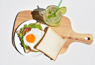 Sandviçli sağlıklı kahvaltı yemeği, kızarmış yumurta, domates dilimi, salata, nane yaprağı, mason kavanozu bardak limonata, ahşap kesim tahtası, beyaz arka plan, güneşli, abur cubur, kilo kaybı, organik, basitlik, vegan, burger, düşük yağ oranı, spor yemeği.