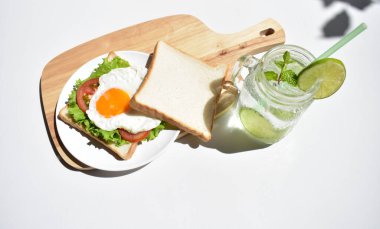 Sandviçli sağlıklı kahvaltı yemeği, kızarmış yumurta, domates dilimi, salata, nane yaprağı, mason kavanozu bardak limonata, ahşap kesim tahtası, beyaz arka plan, güneşli, abur cubur, kilo kaybı, organik, basitlik, vegan, burger, düşük yağ oranı, spor yemeği.