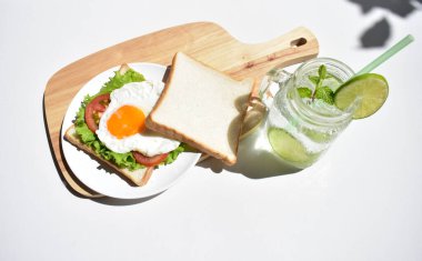 Sandviçli sağlıklı kahvaltı yemeği, kızarmış yumurta, domates dilimi, salata, nane yaprağı, mason kavanozu bardak limonata, ahşap kesim tahtası, beyaz arka plan, güneşli, abur cubur, kilo kaybı, organik, basitlik, vegan, burger, düşük yağ oranı, spor yemeği.